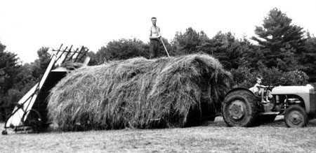 child driving dractor pulling hay wagon with man on top with hay loader trailing behind