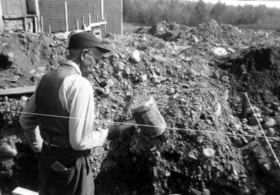 George Hitchcock with pipe hefting mallet in ditch at construction site cris-crossed with string